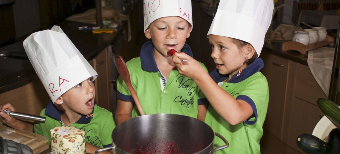 Drei Kinder in Kochmützen und grünen T-Shirts probieren Zutaten während eines Kochkurses im Familienhotel Hinteregger