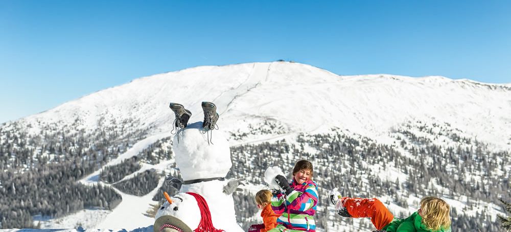 Kinder, die einen Schneemann bauen, während ein anderer Schneemann kopfüber im Schnee steckt, vor einer schneebedeckten Berglandschaft